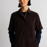A model wearing a dark brown corduroy short-sleeve collared pullover over a blue and black striped long-sleeve shirt, set against a plain light gray background.