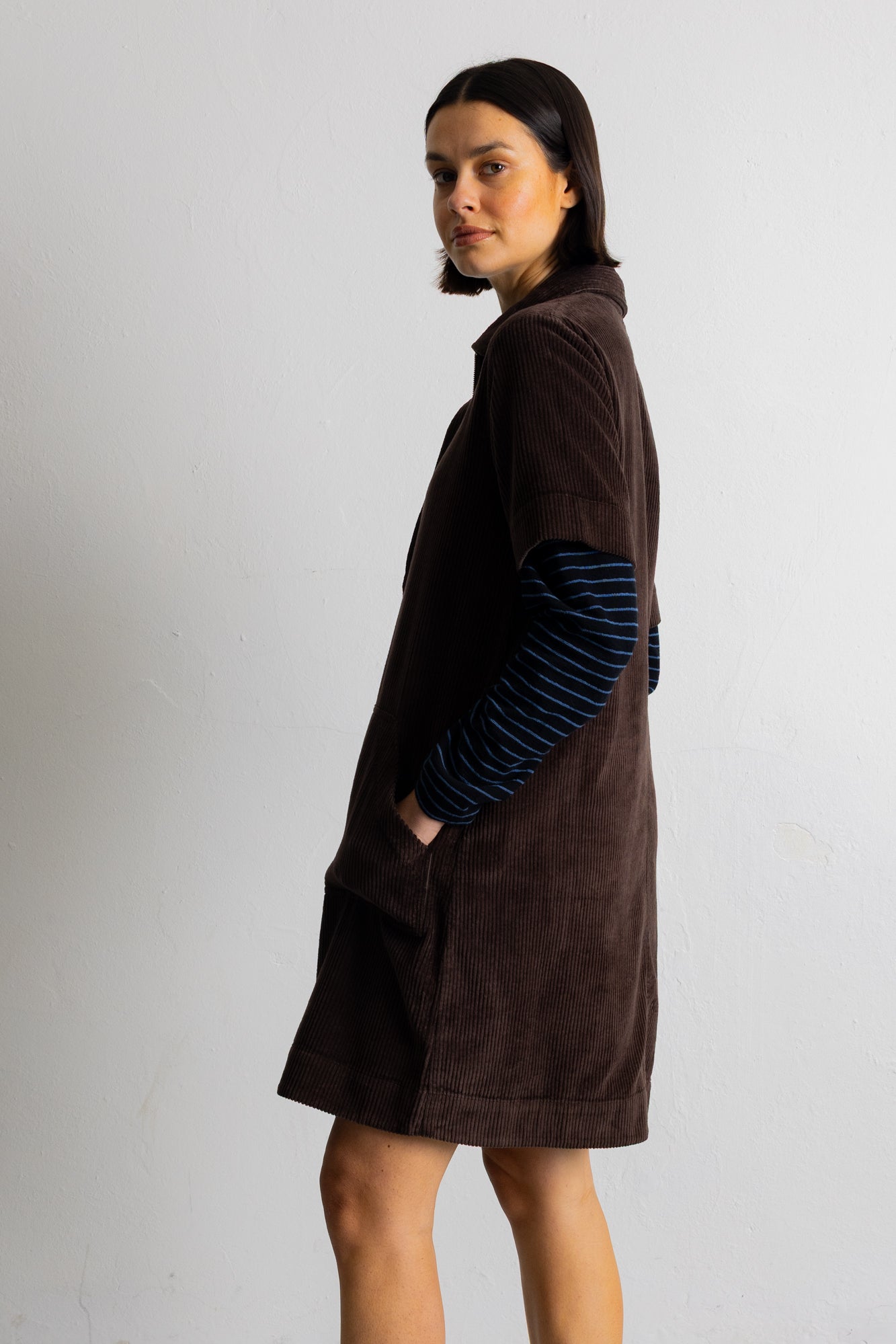 A model wearing a dark brown corduroy short-sleeve collared pullover over a blue and black striped long-sleeve shirt, set against a plain light gray background.