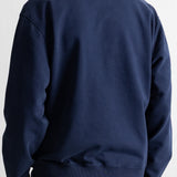 A person shown from the back wearing a navy crewneck sweatshirt and brown pants against a white wall.