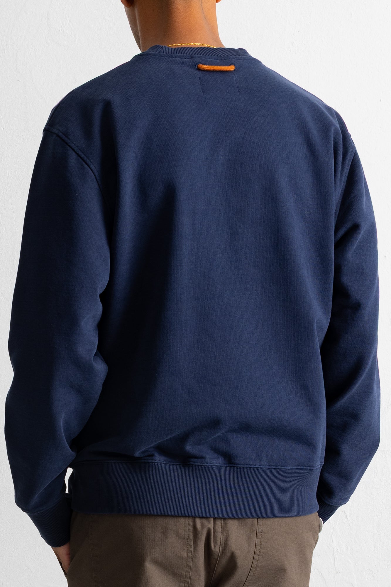 A person shown from the back wearing a navy crewneck sweatshirt and brown pants against a white wall.