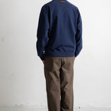 A man viewed from behind wearing a navy blue crewneck sweatshirt and brown wide-leg trousers, standing against a plain white wall.
