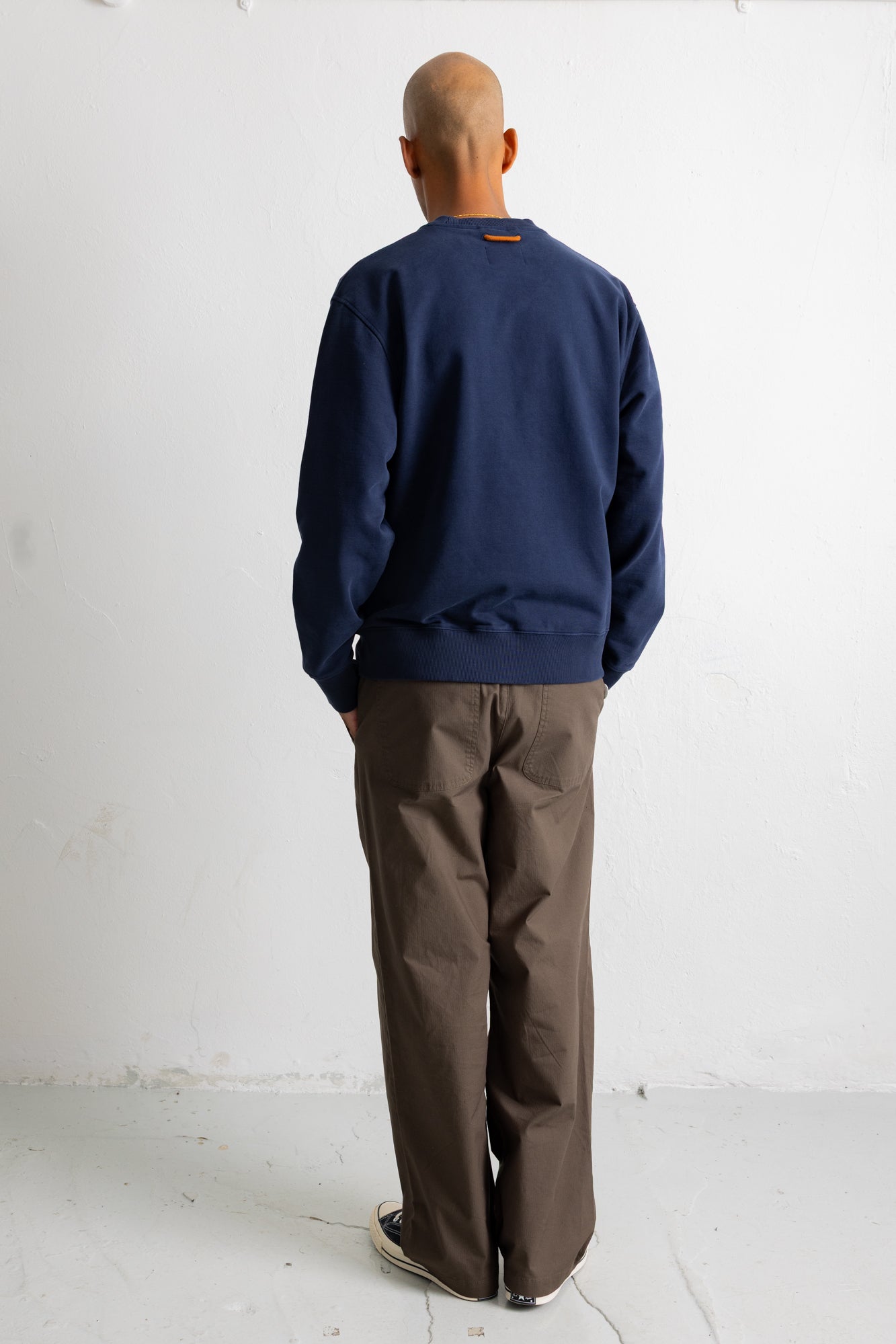 A man viewed from behind wearing a navy blue crewneck sweatshirt and brown wide-leg trousers, standing against a plain white wall.