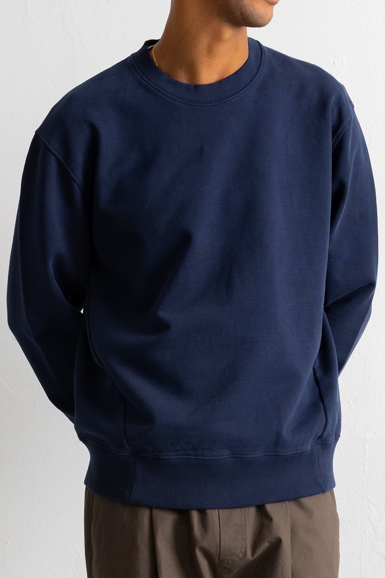 A navy blue crewneck sweatshirt worn by a model, paired with brown pants, against a plain off-white background.