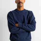 A man with a mustache wearing a navy blue crewneck sweatshirt and brown pants, standing with arms crossed against a plain white wall.