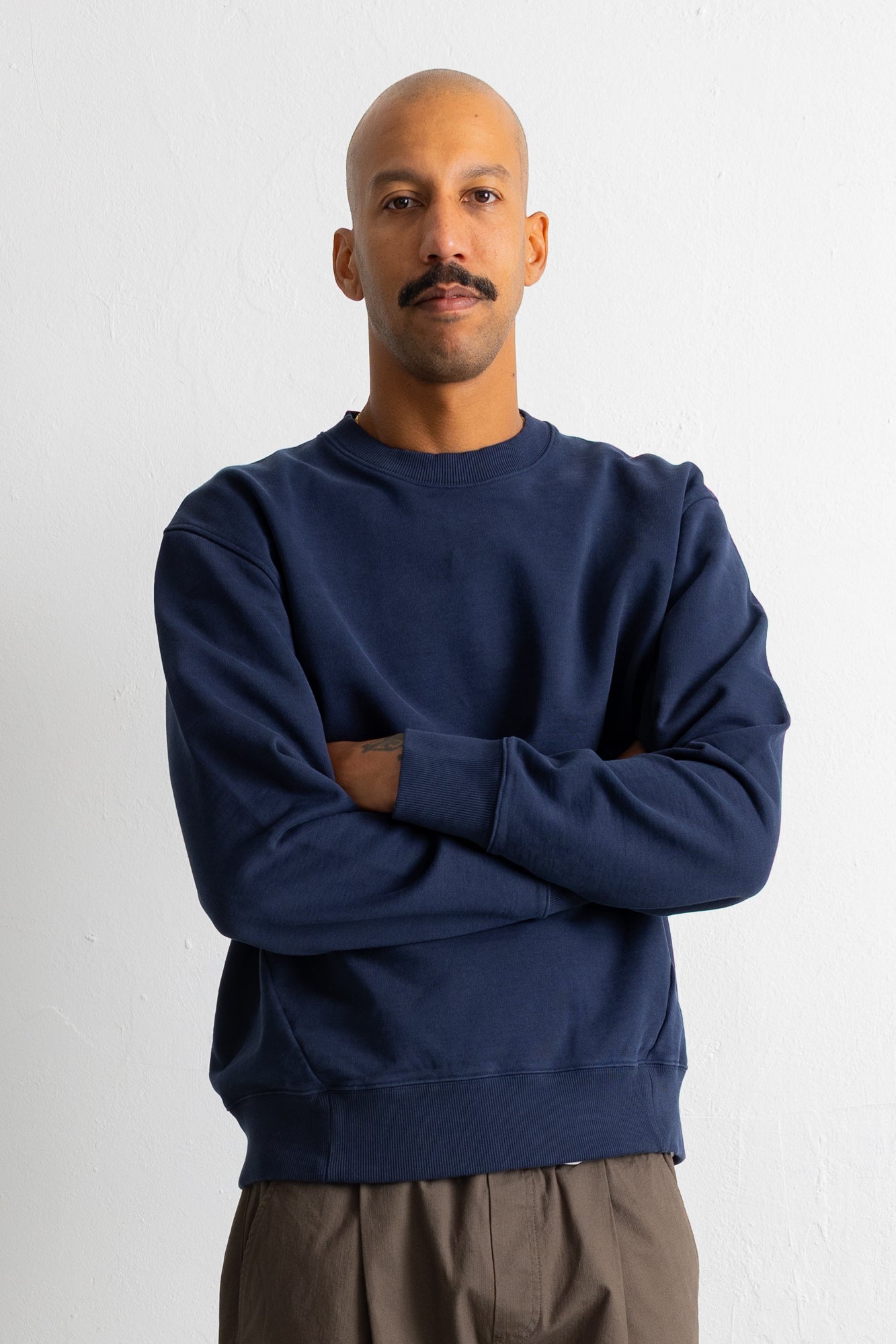 A man with a mustache wearing a navy blue crewneck sweatshirt and brown pants, standing with arms crossed against a plain white wall.