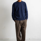 A man wearing a navy blue crewneck sweatshirt, brown trousers, and black canvas sneakers, standing against a plain white wall.