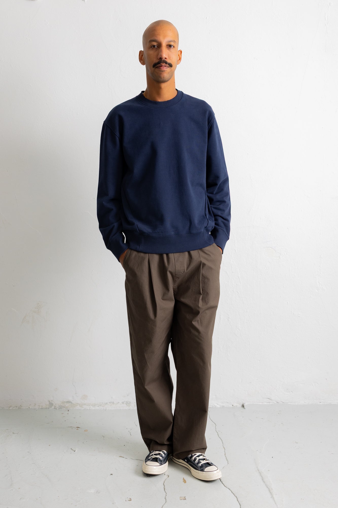 A man wearing a navy blue crewneck sweatshirt, brown trousers, and black canvas sneakers, standing against a plain white wall.