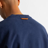 Close-up back view of a person wearing a navy crewneck sweatshirt with a small orange tab detail at the back of the collar, against a plain white wall.