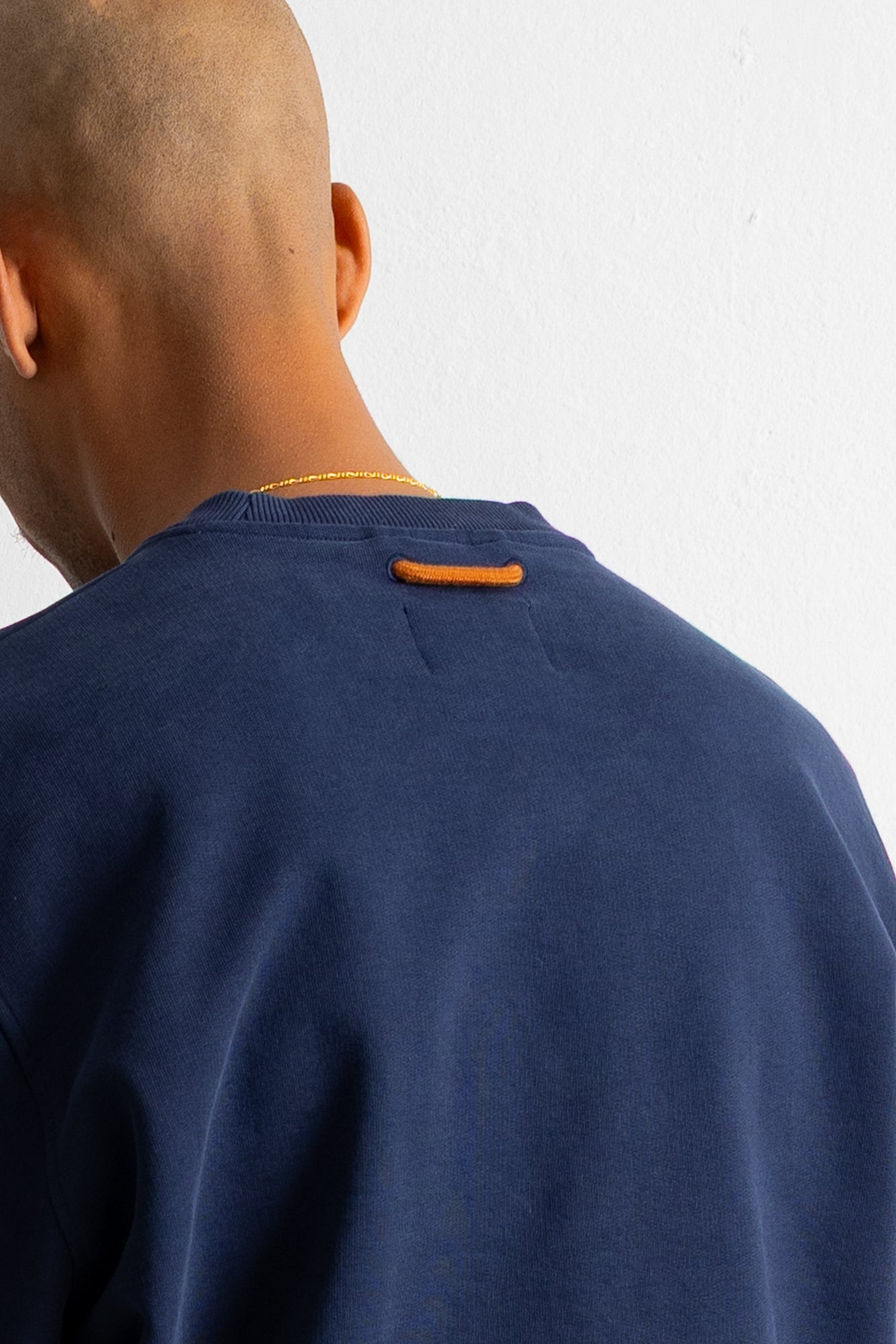 Close-up back view of a person wearing a navy crewneck sweatshirt with a small orange tab detail at the back of the collar, against a plain white wall.