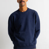 A man wearing a navy blue crewneck sweatshirt and brown pants, standing against a plain white wall.