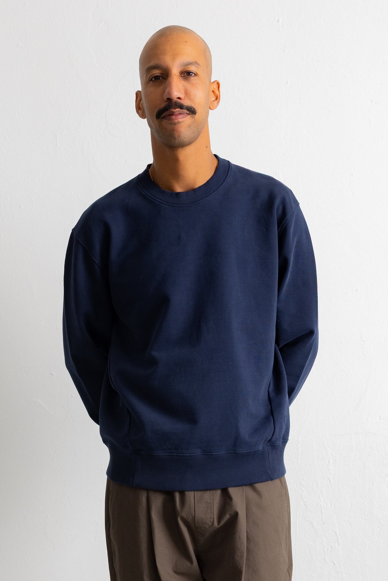 A man wearing a navy blue crewneck sweatshirt and brown pants, standing against a plain white wall.