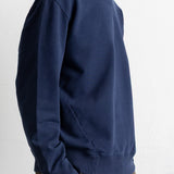 A man wearing a navy blue crewneck sweatshirt and brown trousers, shown in a side profile against a plain white wall.