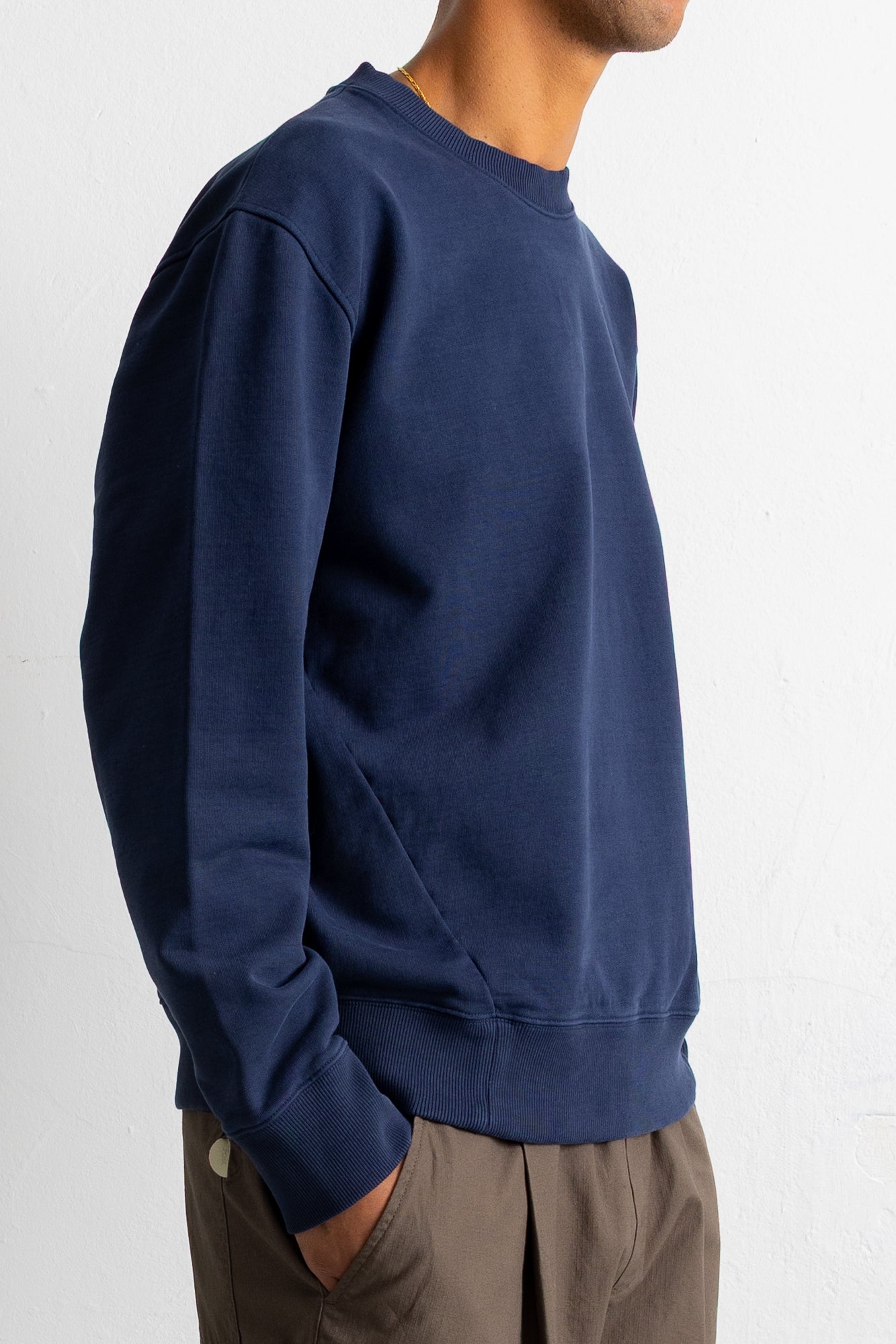 A man wearing a navy blue crewneck sweatshirt and brown trousers, shown in a side profile against a plain white wall.