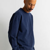 A man wearing a navy blue crewneck sweatshirt and brown trousers, standing against a plain white wall.