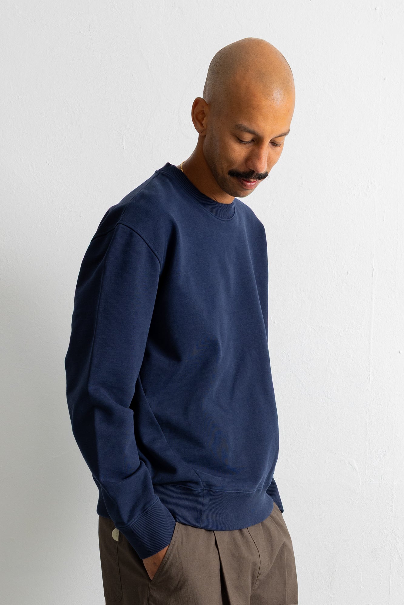 A man wearing a navy blue crewneck sweatshirt and brown trousers, standing against a plain white wall.
