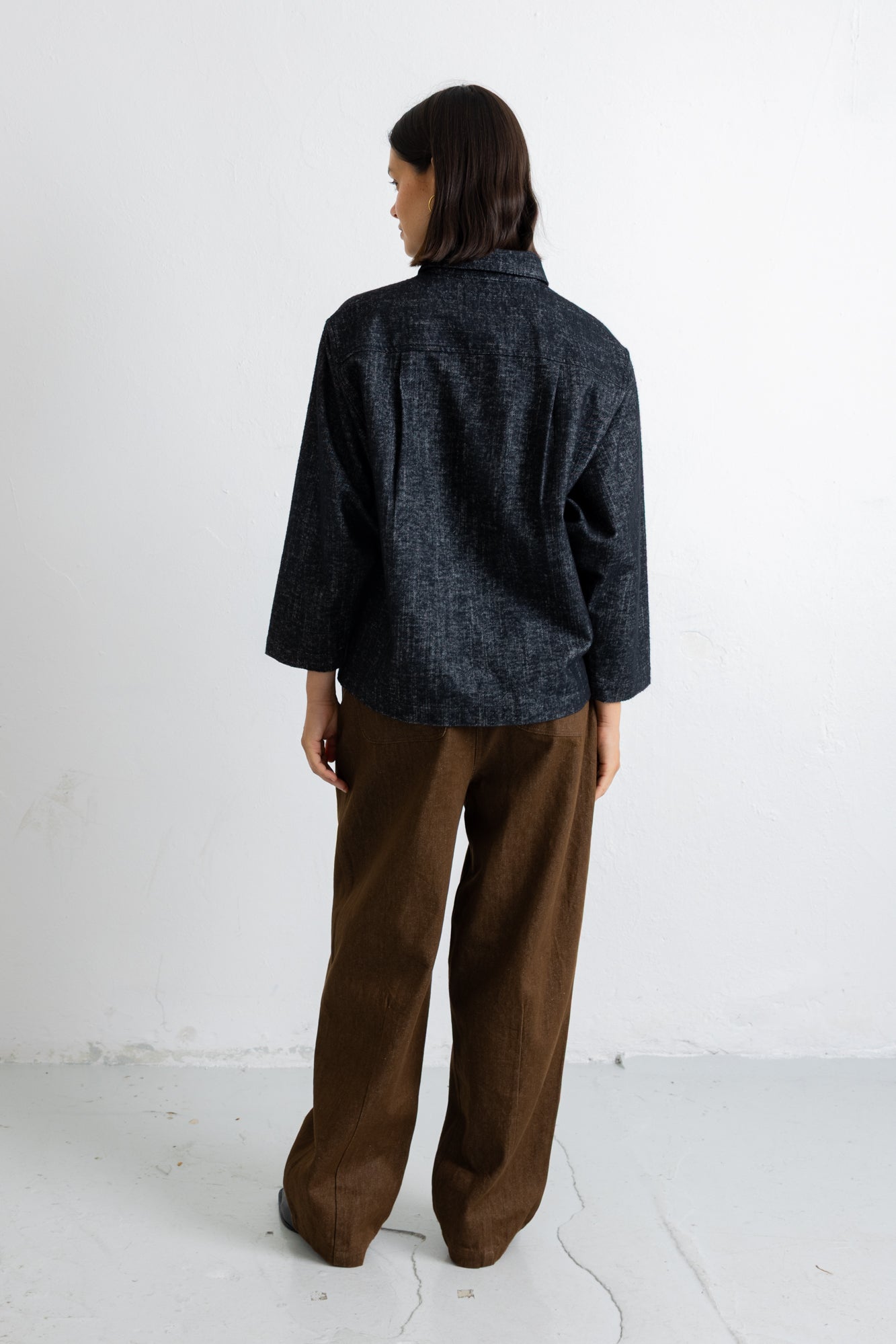 A model shown from the back wearing a dark navy textured long-sleeve shirt and brown wide-leg trousers, standing against a plain white wall.