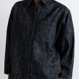 A woman wearing a dark charcoal gray textured button-up shirt with brown pants, shown from the chin down against a plain white wall.
