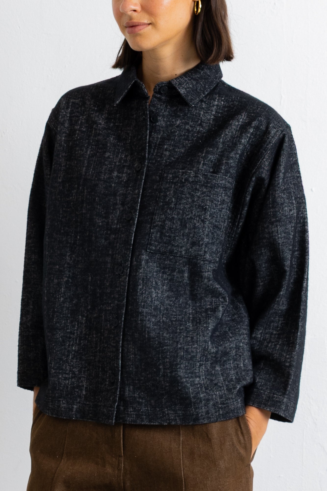 A woman wearing a dark charcoal gray textured button-up shirt with brown pants, shown from the chin down against a plain white wall.