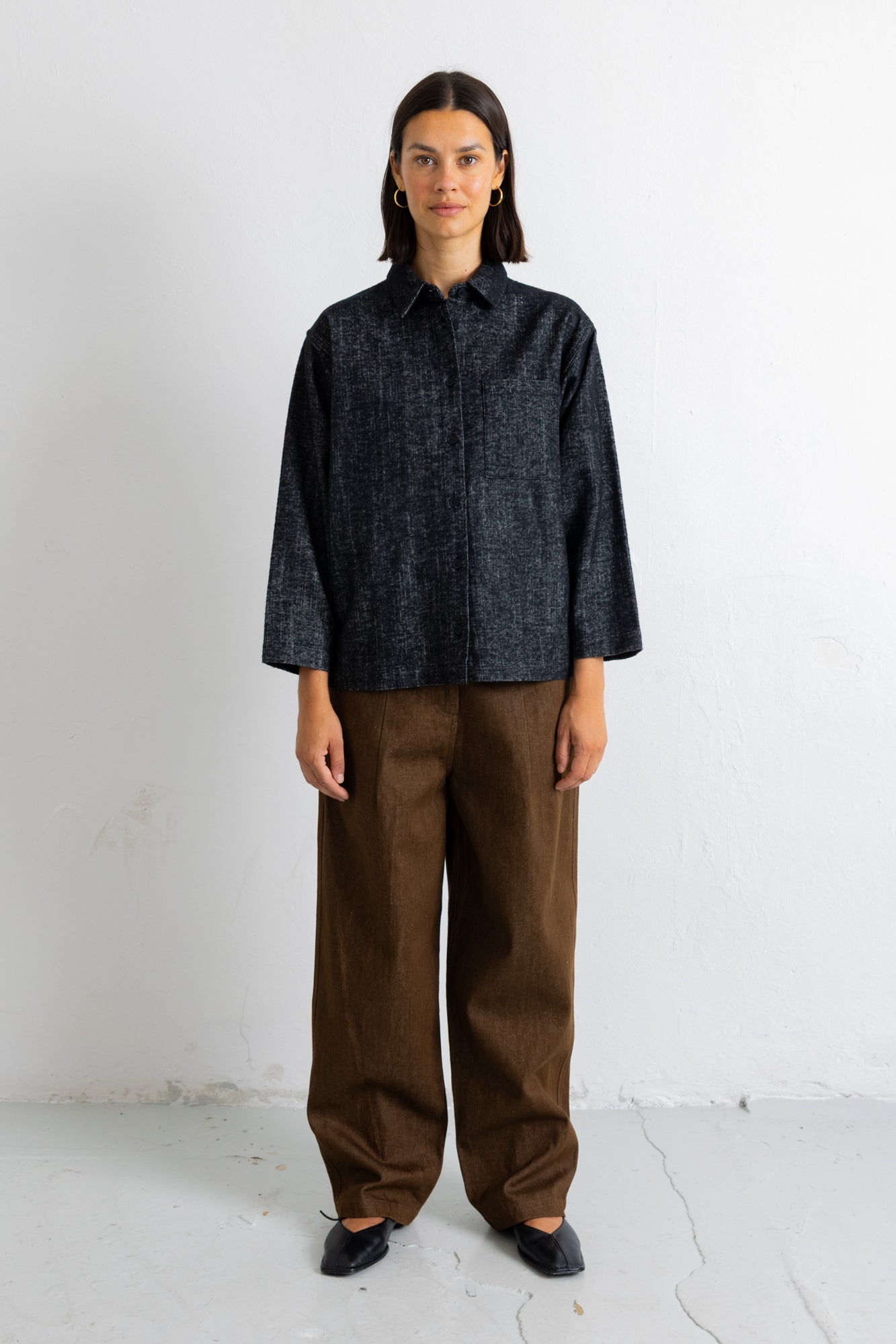 A model stands against a white wall wearing a dark navy textured long-sleeve button-up shirt and wide-leg brown trousers.