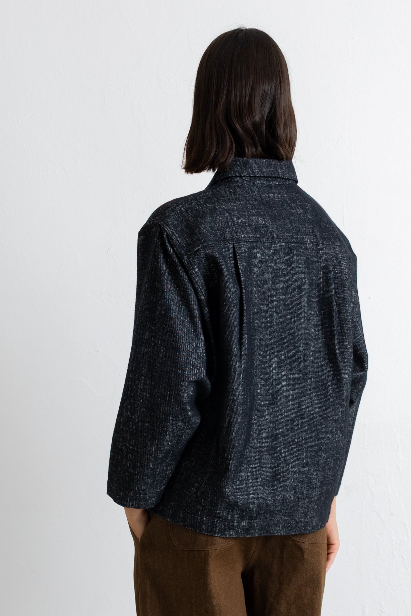 A person shown from the back wearing a dark charcoal textured overshirt and brown pants against a plain white wall.