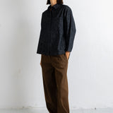 A woman stands in a minimalist studio setting wearing a dark textured button-up shirt and brown wide-leg trousers.