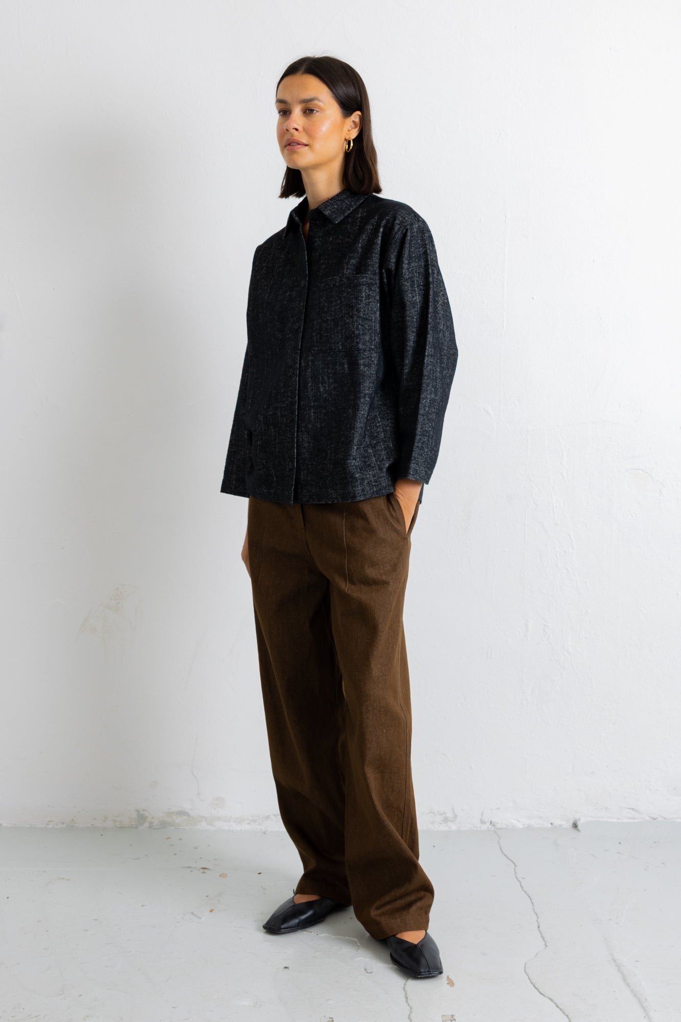 A woman stands in a minimalist studio setting wearing a dark textured button-up shirt and brown wide-leg trousers.