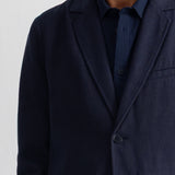 A close-up of a man wearing a dark navy blue corduroy blazer over a matching navy shirt, against a plain white wall.