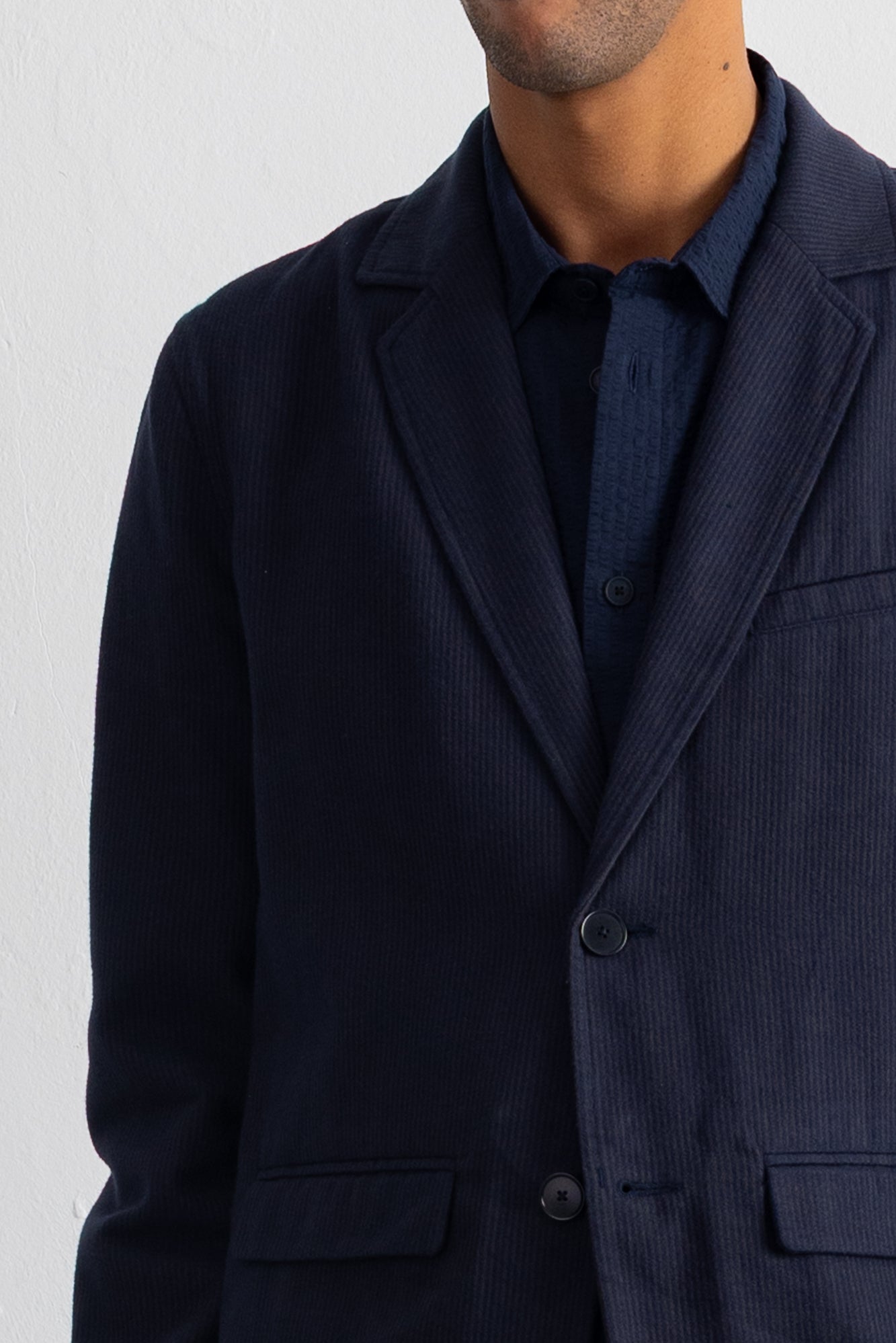 A close-up of a man wearing a dark navy blue corduroy blazer over a matching navy shirt, against a plain white wall.