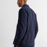 A man shown from the back wearing a dark navy blue jacket and matching trousers against a plain white wall.