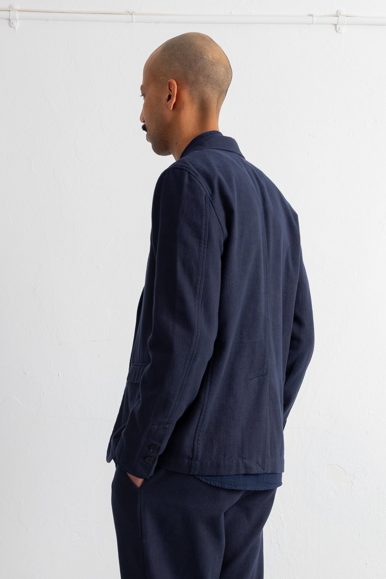 A man shown from the back wearing a dark navy blue jacket and matching trousers against a plain white wall.