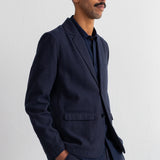 A man wearing a dark navy blue blazer and matching trousers over a dark shirt, standing against a plain light gray wall.