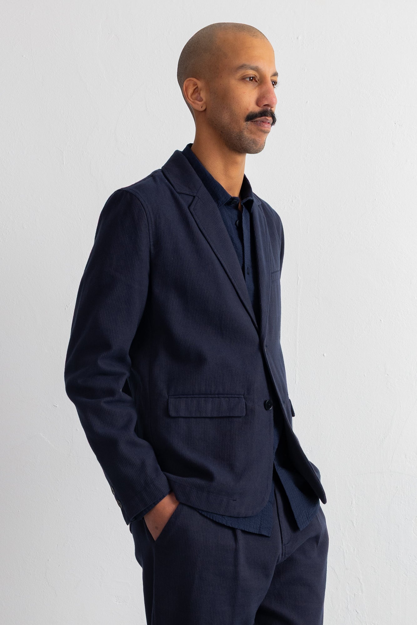 A man wearing a dark navy blue blazer and matching trousers over a dark shirt, standing against a plain light gray wall.