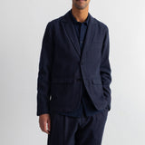 A man wearing a dark navy blue blazer and matching trousers over a dark shirt, standing against a plain white wall.