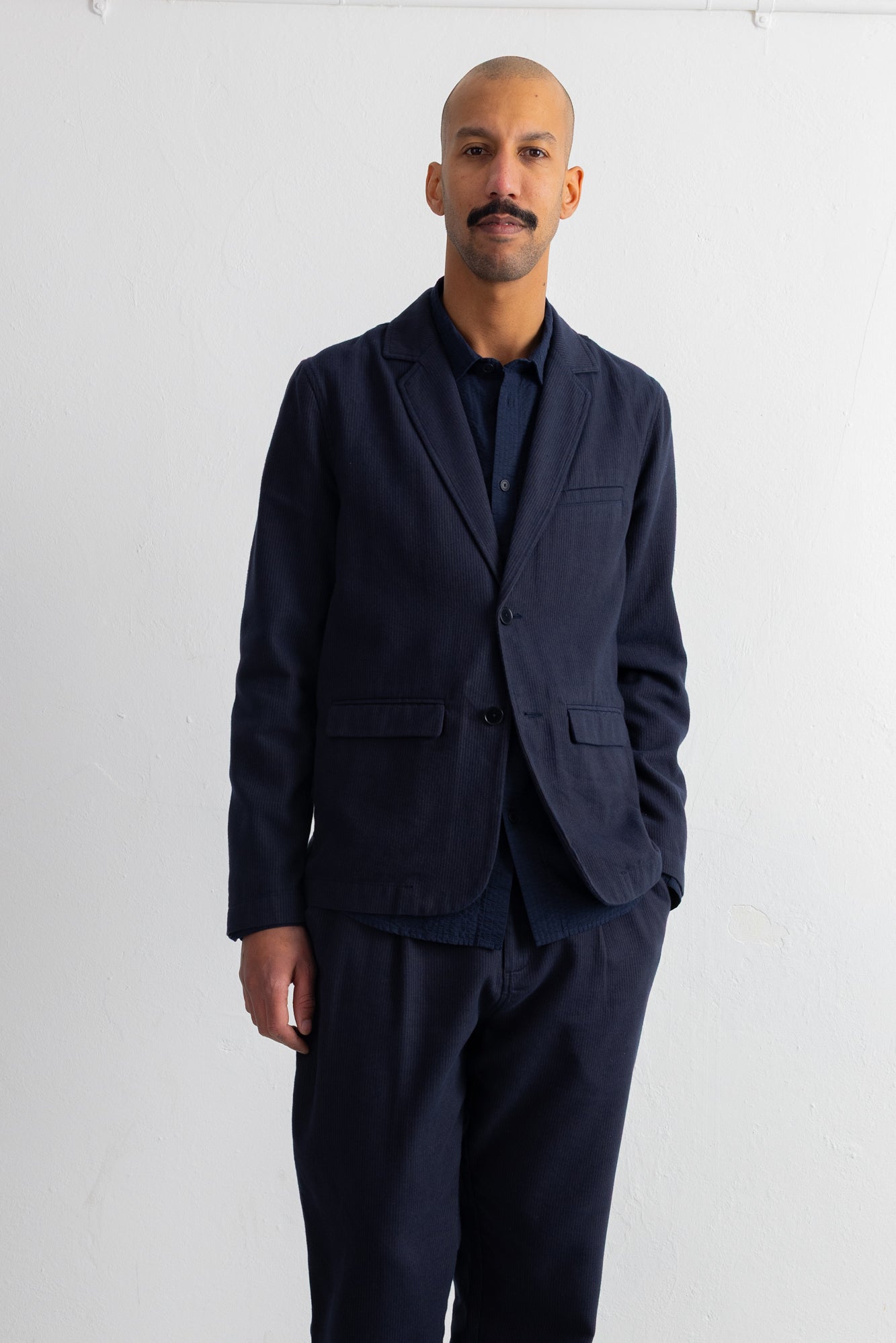 A man wearing a dark navy blue blazer and matching trousers over a dark shirt, standing against a plain white wall.