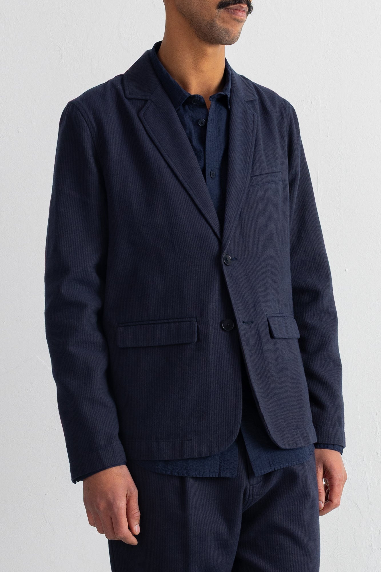 A man wearing a dark navy blue blazer over a matching navy shirt and trousers, shown from the chin down against a plain white background.