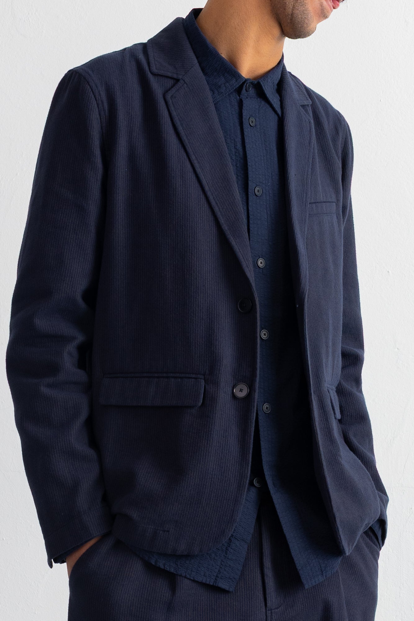 A man wearing a dark navy blue corduroy blazer over a matching navy shirt, shown from the neck down against a plain light background.