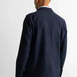A man viewed from behind wearing a dark navy blue blazer and matching trousers against a plain white background.