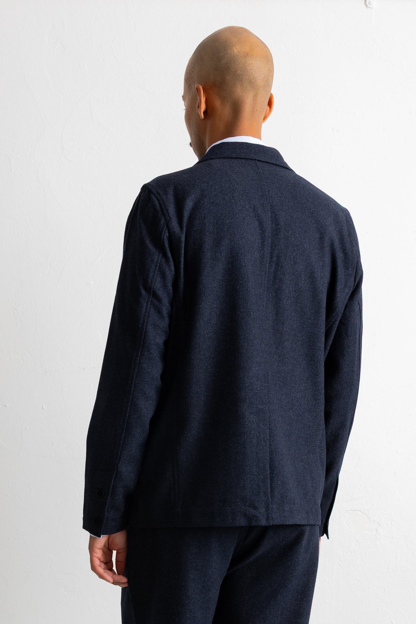 A man viewed from behind wearing a dark navy blue blazer and matching trousers against a plain white background.