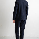 A man viewed from behind wearing a dark navy blue suit, consisting of a jacket and trousers, paired with dark brown leather shoes, standing against a plain white studio background.
