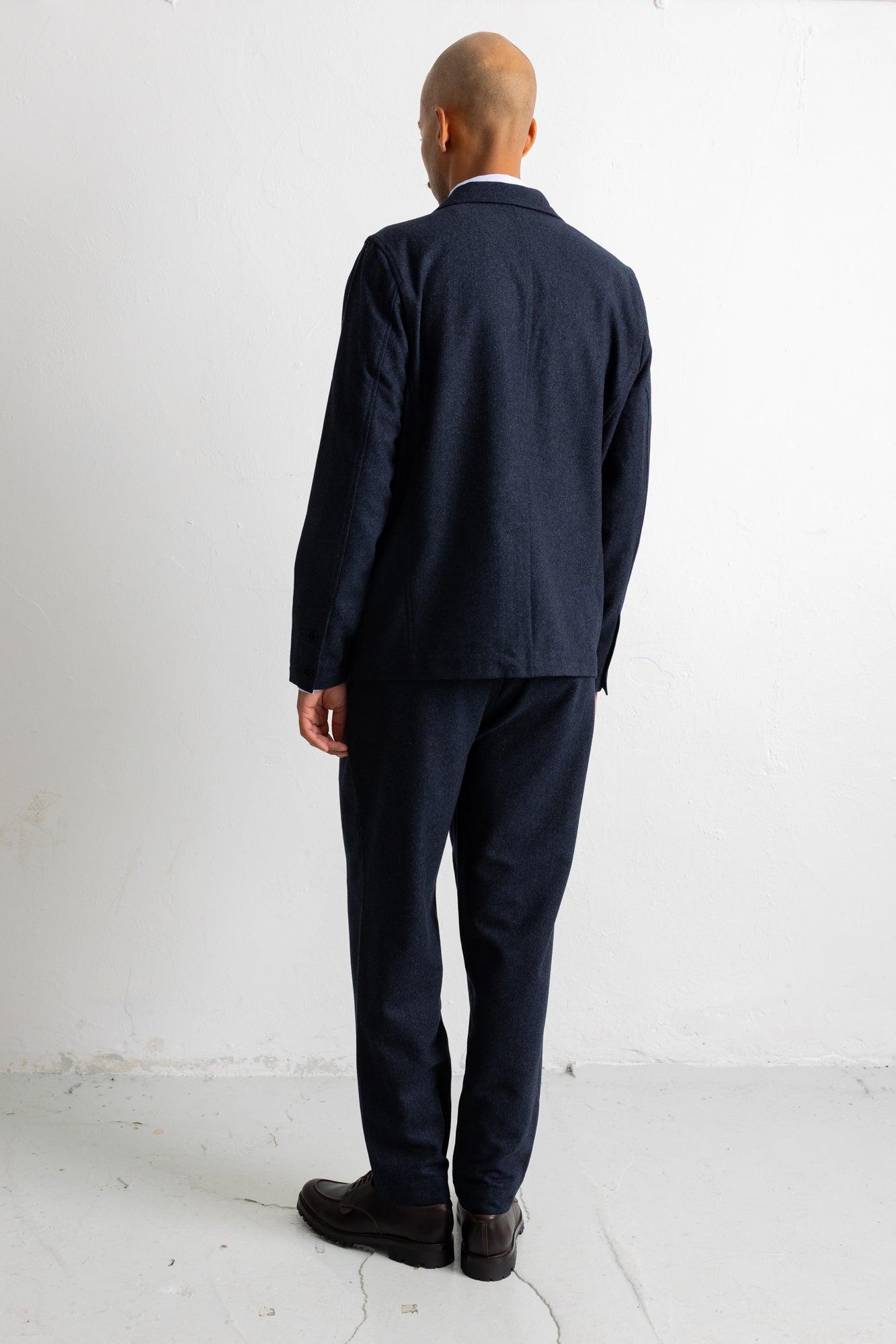A man viewed from behind wearing a dark navy blue suit, consisting of a jacket and trousers, paired with dark brown leather shoes, standing against a plain white studio background.