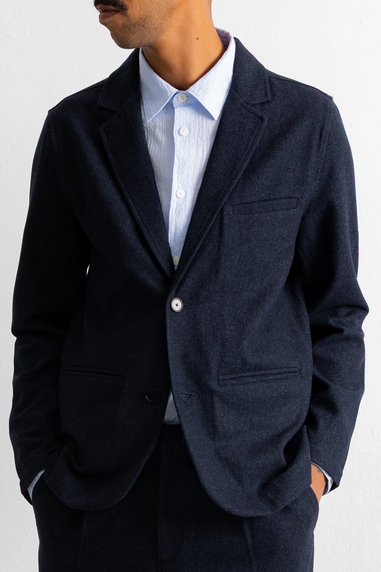 A man wearing a dark navy blue textured blazer over a light blue striped dress shirt, shown from the chin down against a plain white background.