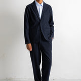 A man wearing a dark navy blue suit with a light blue dress shirt and dark brown leather shoes, standing against a white studio background.