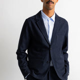 A man wearing a dark navy blue textured blazer over a light blue dress shirt, standing against a plain light gray background.