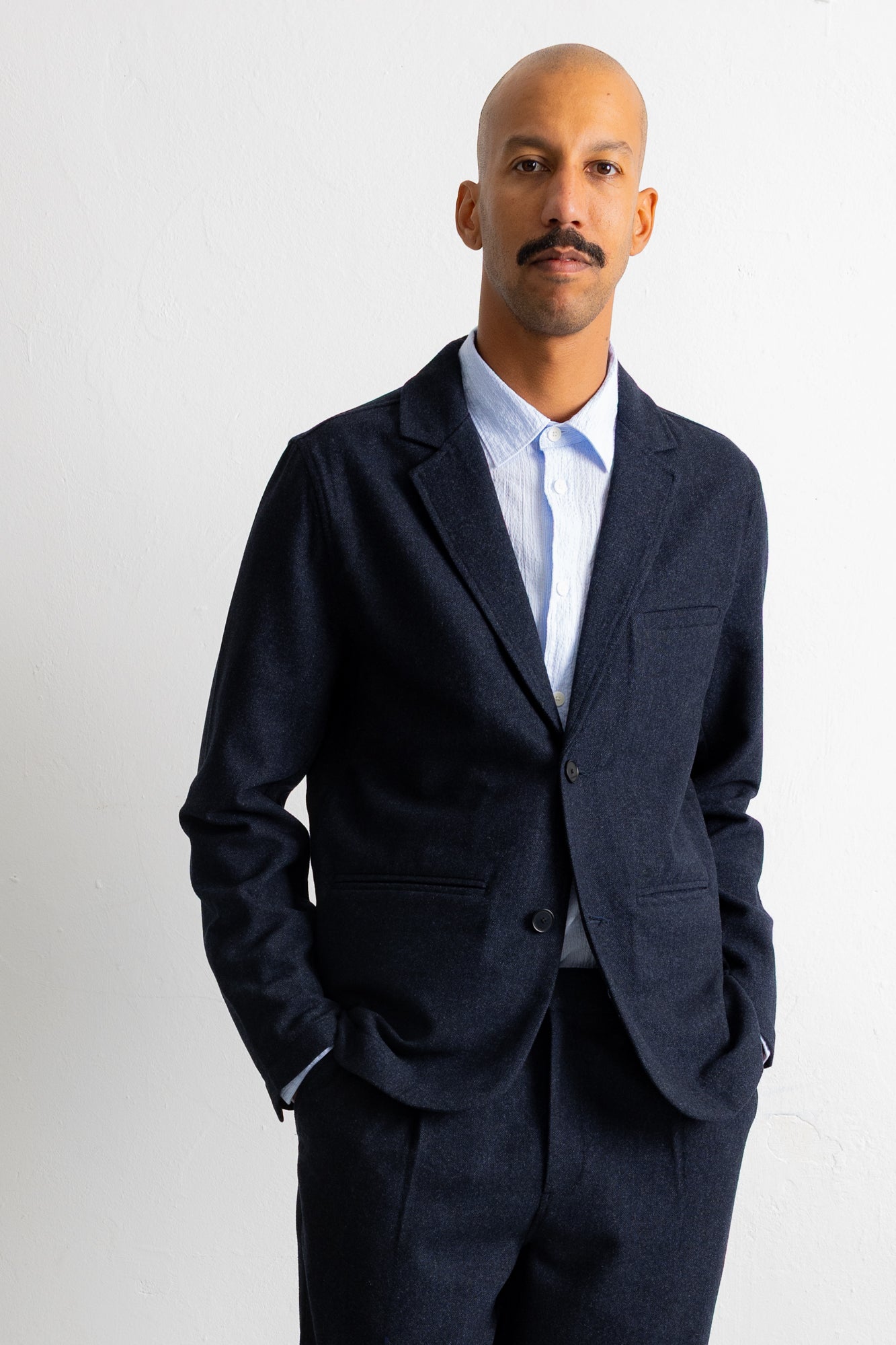 A man wearing a dark navy blue textured blazer over a light blue dress shirt, standing against a plain light gray background.