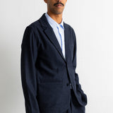 A man wearing a dark navy blue suit jacket and matching trousers over a light blue collared shirt, standing against a plain white wall.