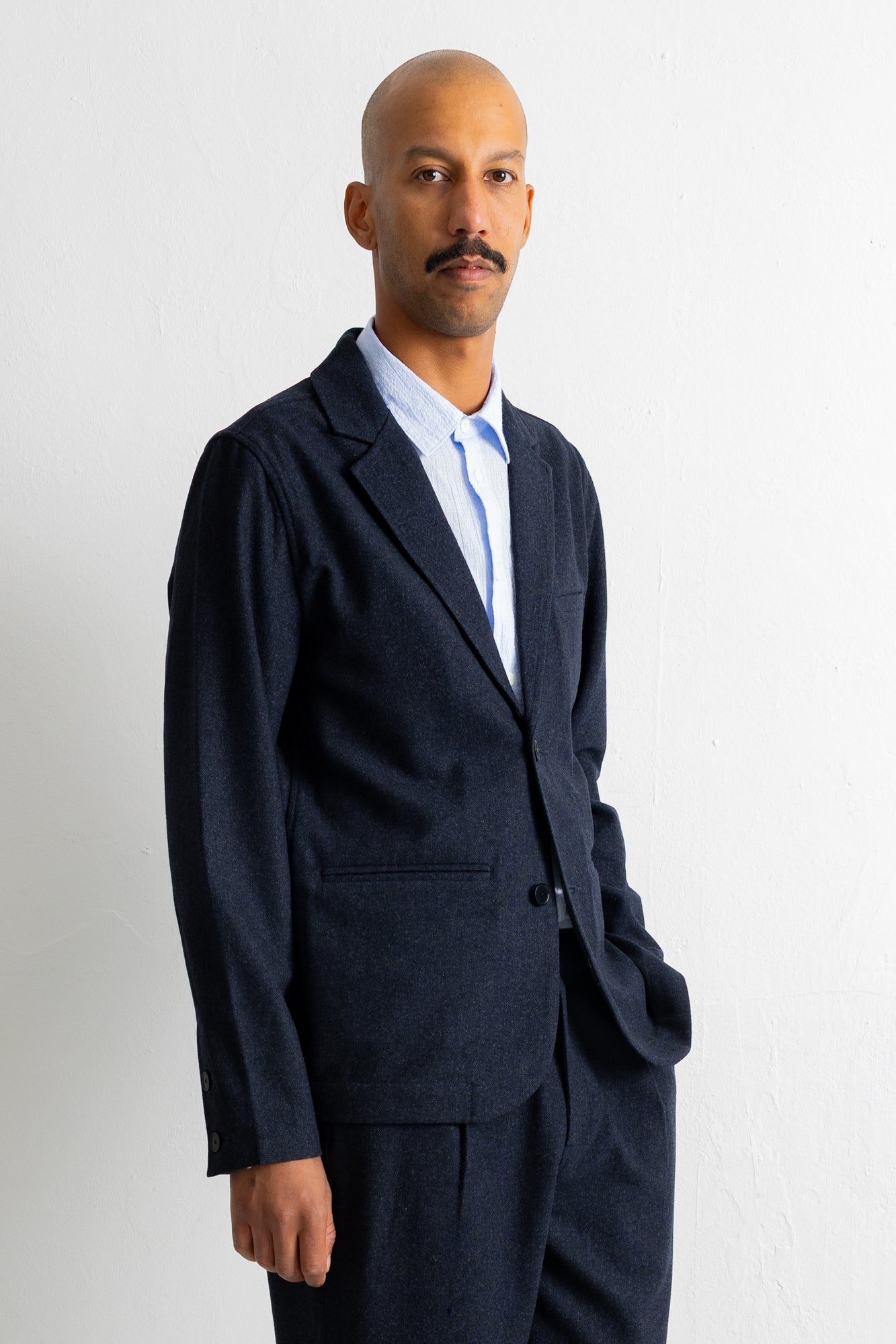A man wearing a dark navy blue suit jacket and matching trousers over a light blue collared shirt, standing against a plain white wall.