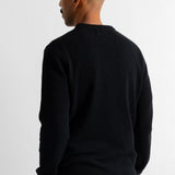 A man viewed from the back wearing a black long-sleeve knit sweater and dark blue corduroy trousers, standing against a plain white wall.