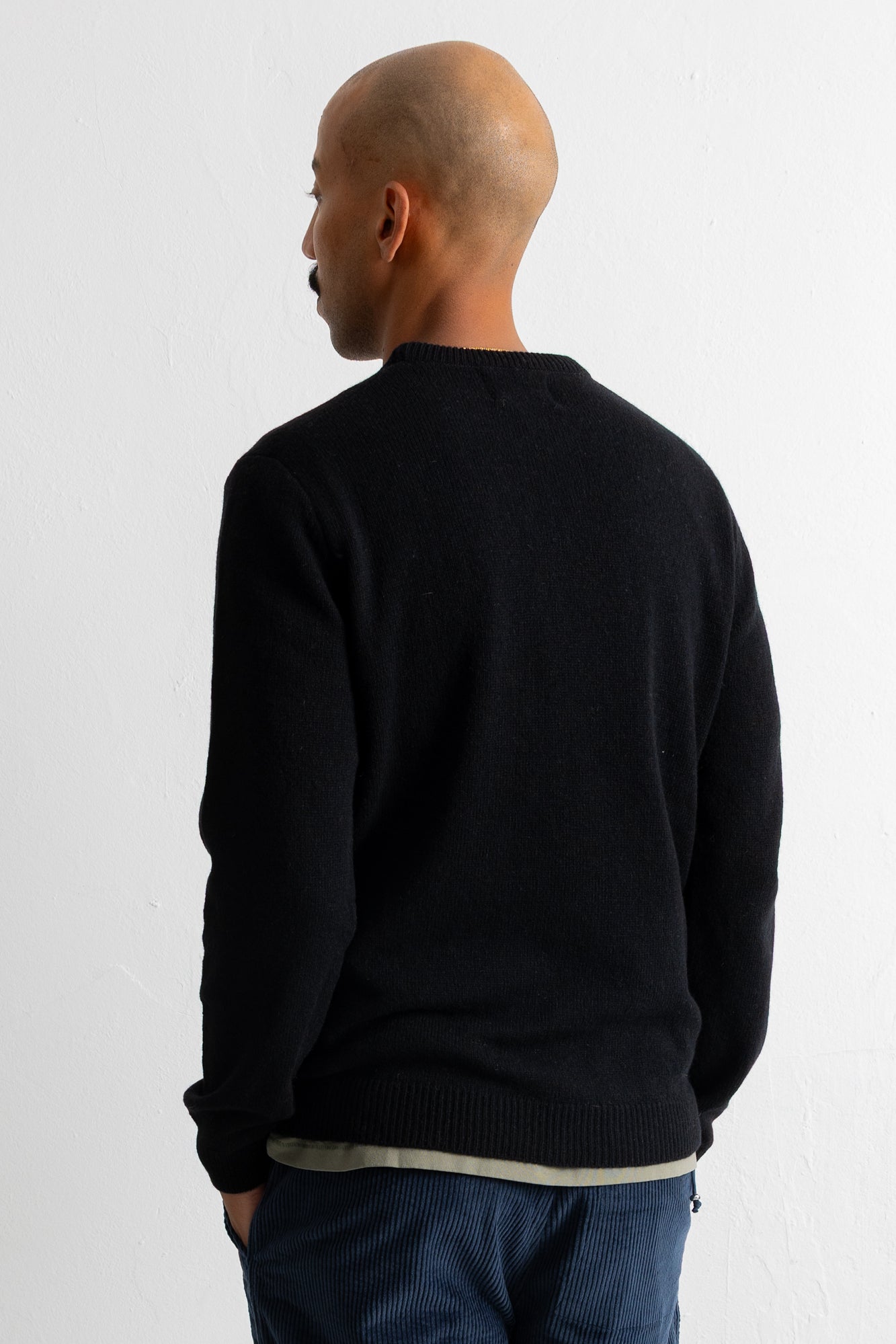 A man viewed from the back wearing a black long-sleeve knit sweater and dark blue corduroy trousers, standing against a plain white wall.