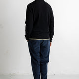 A man viewed from behind wearing a black crewneck sweater, dark blue jeans, and black canvas sneakers, standing against a plain white studio wall.