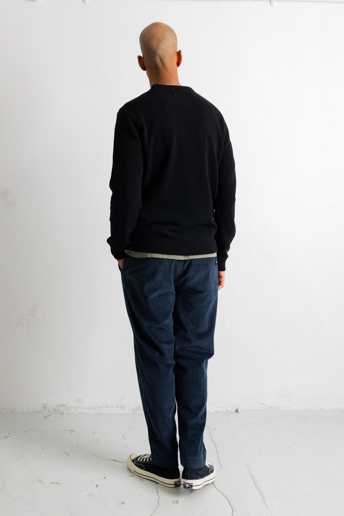 A man viewed from behind wearing a black crewneck sweater, dark blue jeans, and black canvas sneakers, standing against a plain white studio wall.